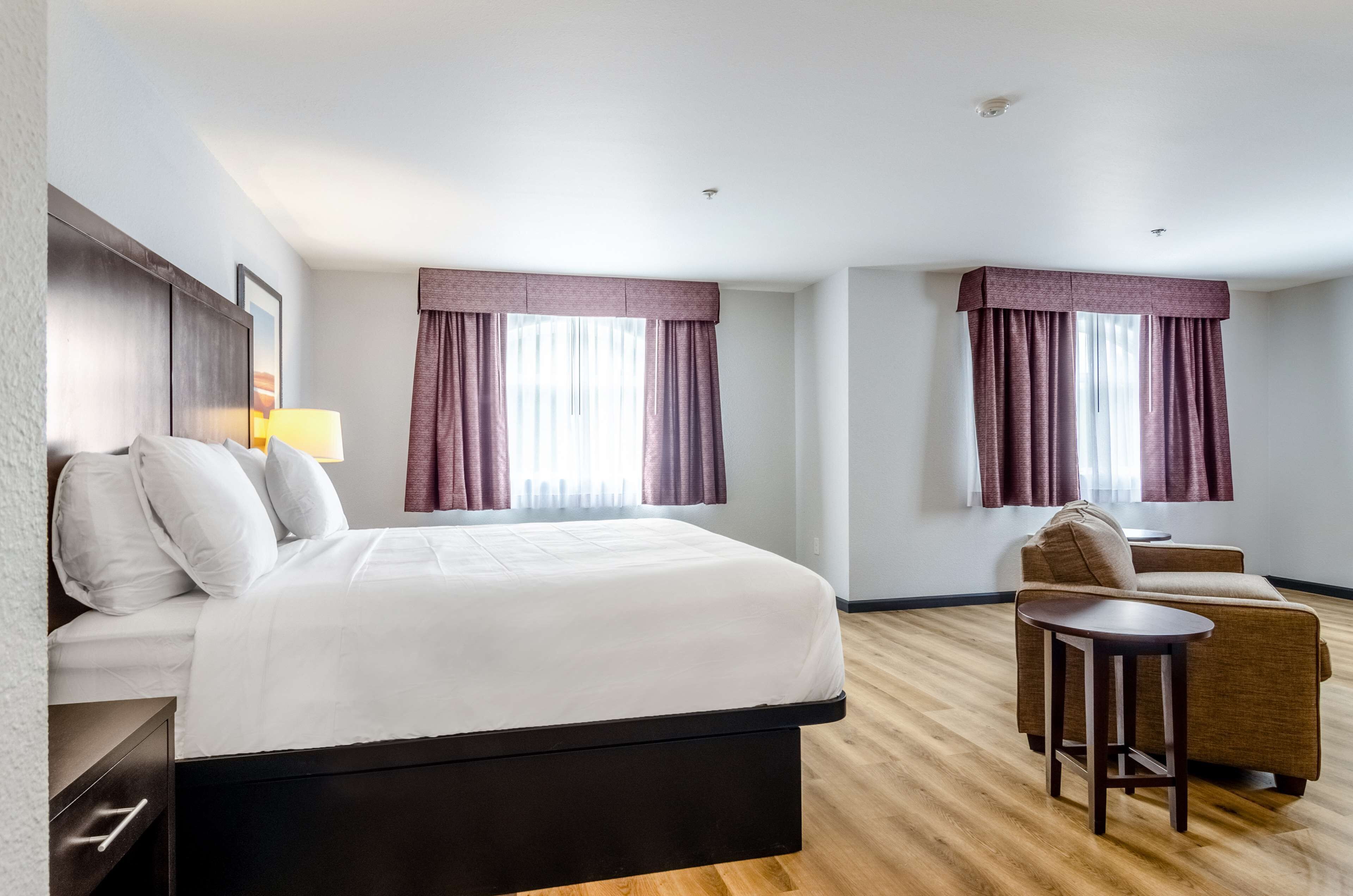 A hotel room with a bed, a sofa, a small table, and two windows with curtains.