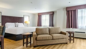 Une chambre d’hôtel avec un lit, un canapé, deux tables de chevet, une lampe et de grandes fenêtres avec des rideaux.