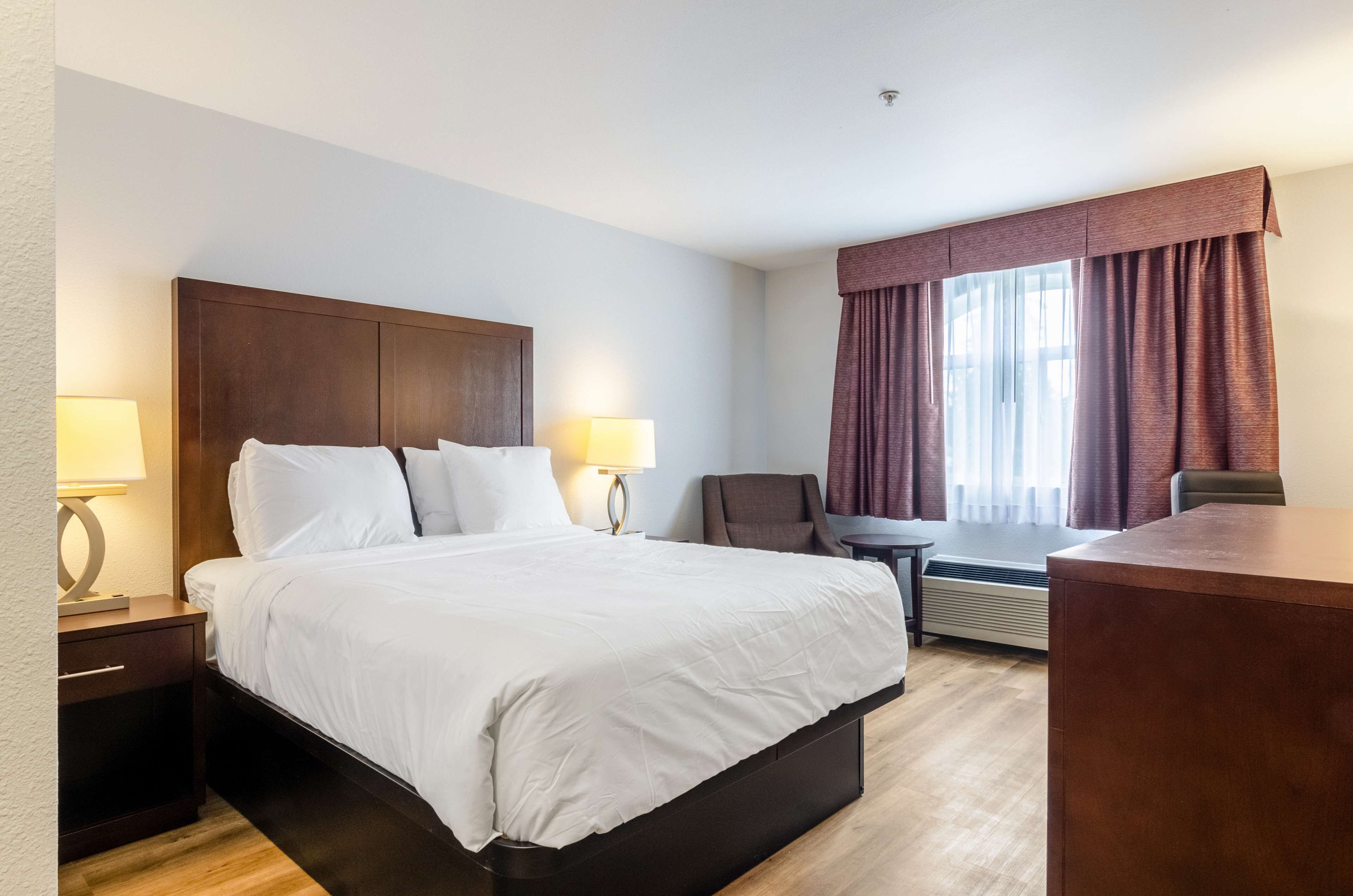 A hotel room with a large bed, two bedside tables with lamps, a chair, a desk, and a window with curtains.