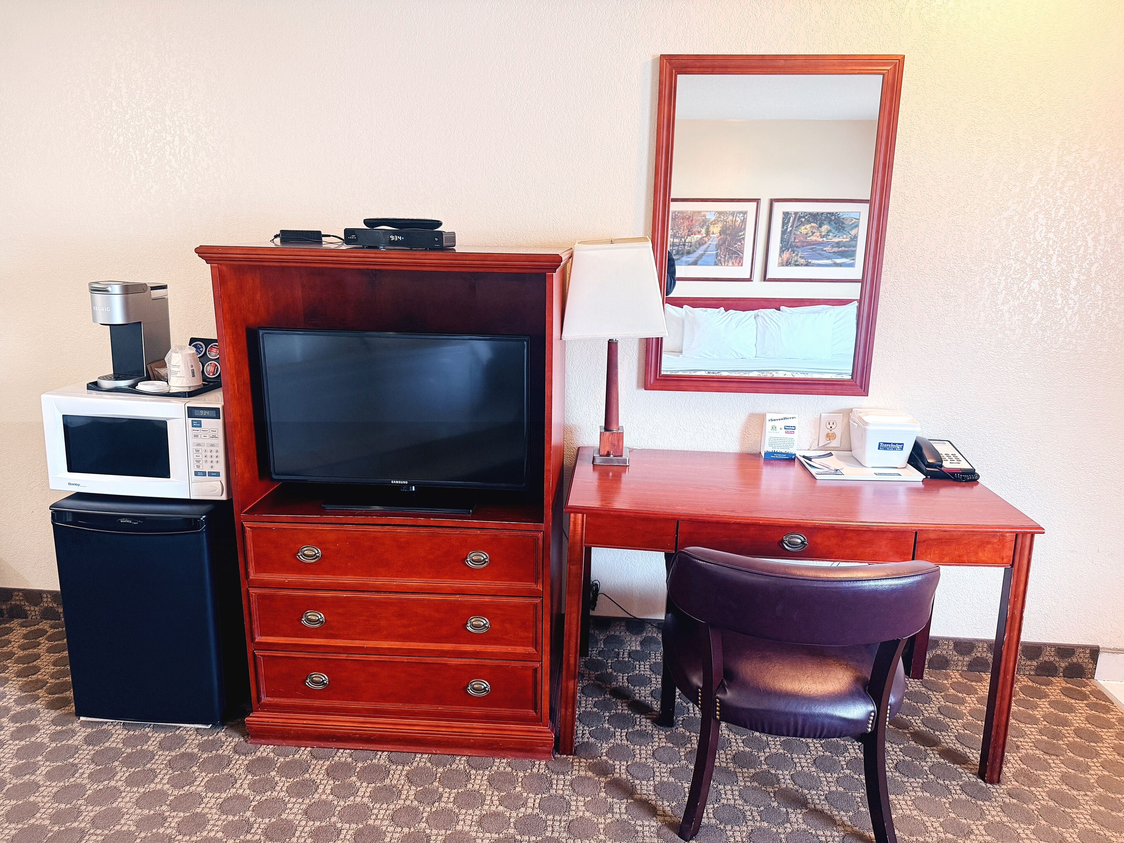 A hotel room with a wooden desk, a television, a microwave, a coffee maker, a refrigerator, a chair, and a mirror.