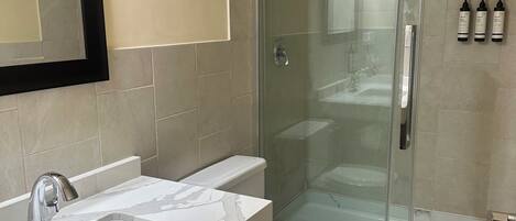 Une salle de bain moderne avec une douche à parois de verre, un lavabo blanc doté d’un plan de toilette en marbre et des toilettes.