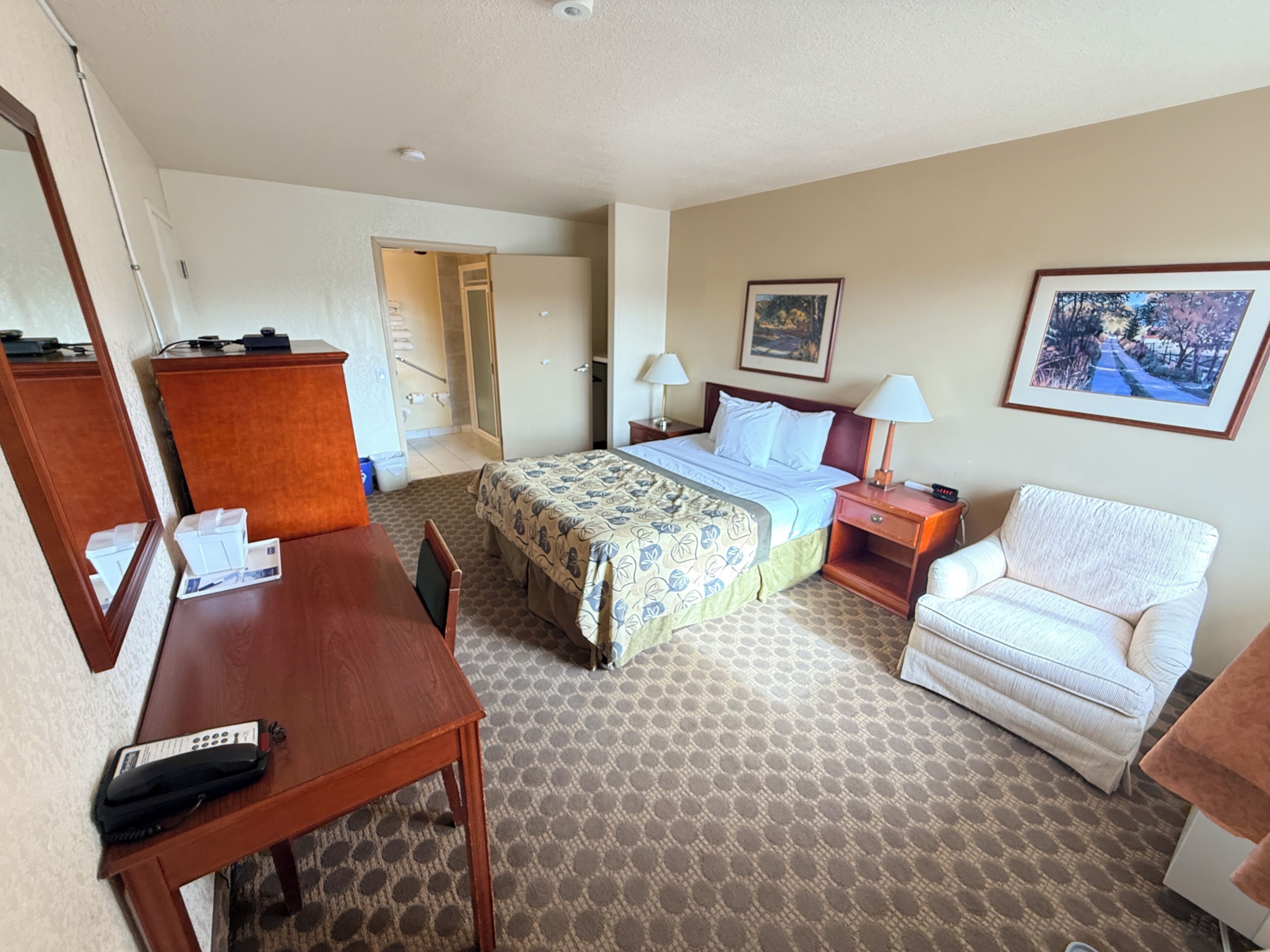 Une chambre d’hôtel comprenant un lit, un bureau, une chaise, une table de chevet et une porte donnant sur un couloir.