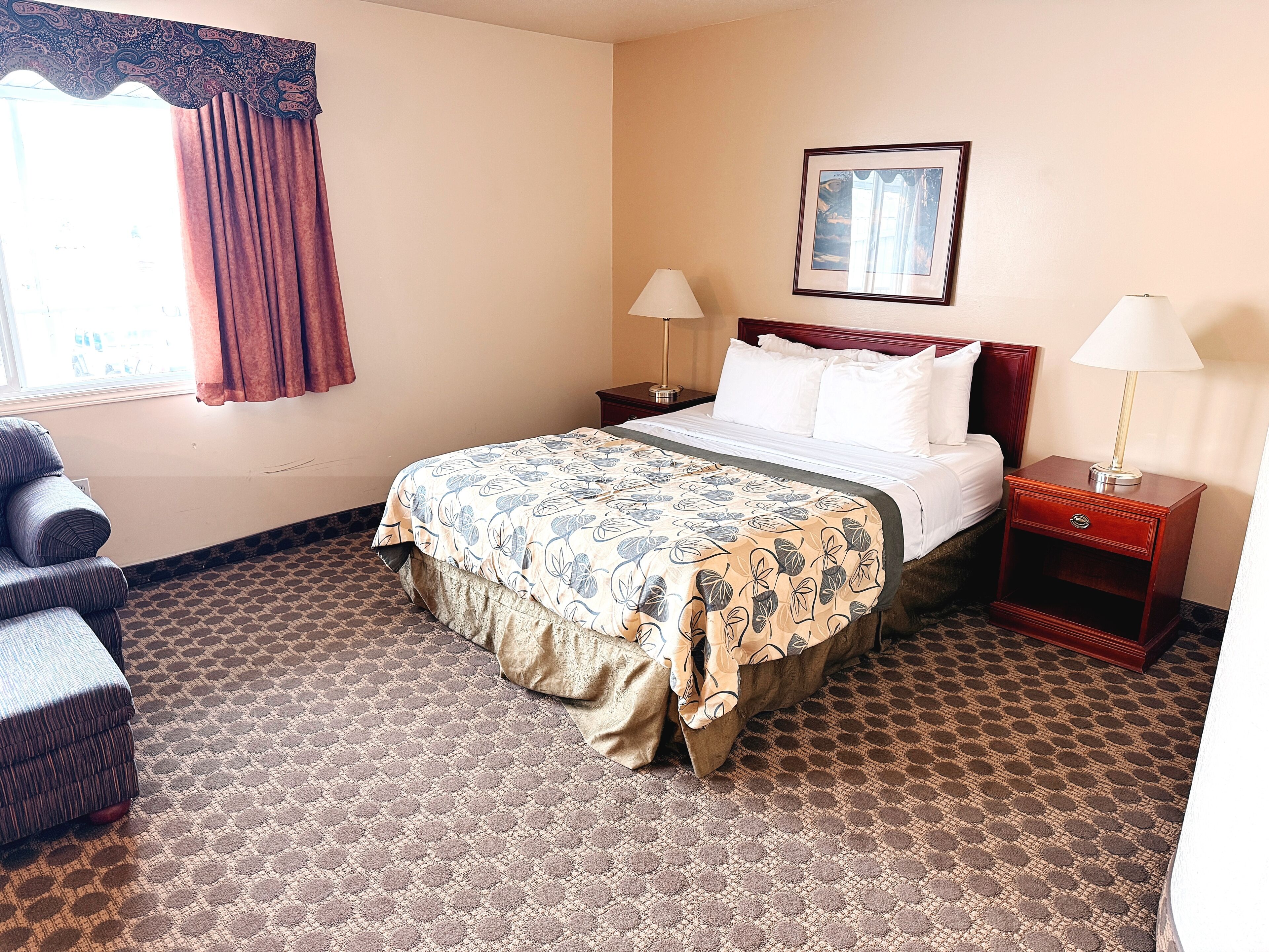 A hotel room with a bed, two nightstands, a chair, a window with curtains, and a painting on the wall.