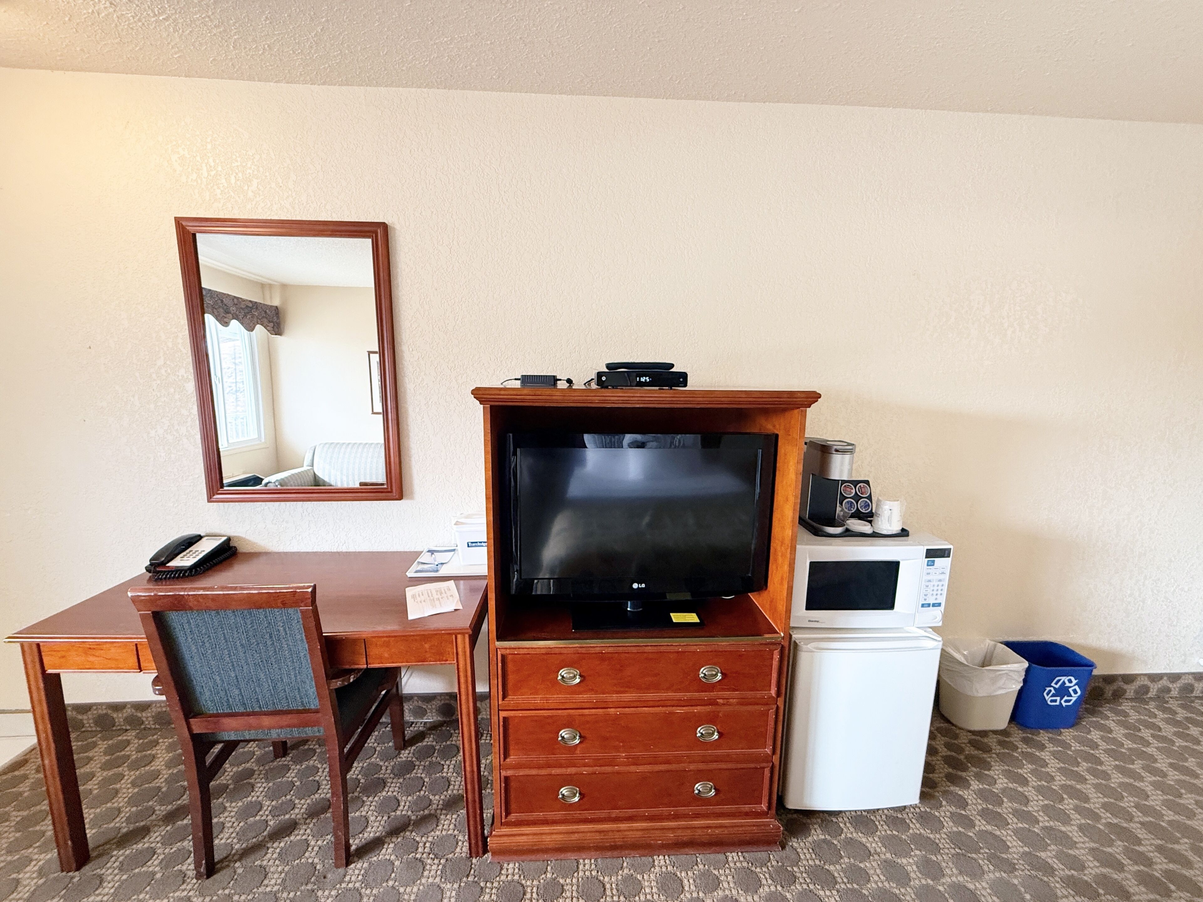 Habitación de hotel con escritorio de madera, televisión, microondas, refrigerador pequeño, bote de basura y espejo.