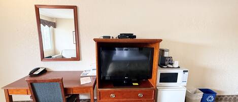 Habitación de hotel con escritorio de madera, televisión, microondas, refrigerador pequeño, bote de basura y espejo.