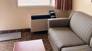 Una sala de estar con sofá, una mesa, equipo de aire acondicionado, ventana con cortinas y una alfombra estampada.
