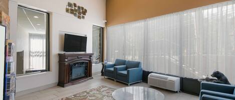 Una sala de estar con chimenea, televisor, sofá azul, mesa de centro de vidrio y un ventanal con cortinas translúcidas.