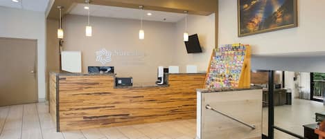 A hotel reception area with a wooden reception desk, a mounted television, and a wall-mounted sign for SureStay Plus Hotel by Best Western.