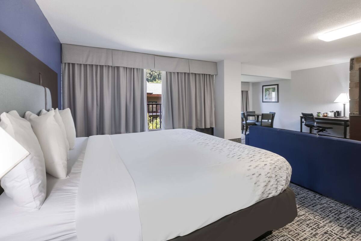 Une chambre d’hôtel avec un grand lit, un bureau et une chaise. Il y a une fenêtre avec des rideaux, et on aperçoit un balcon à travers la fenêtre.