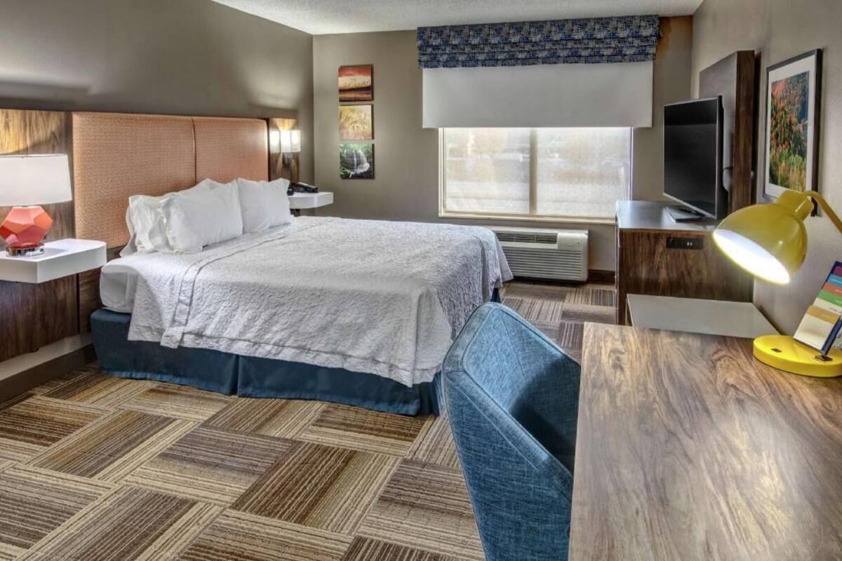 A hotel room with a bed, bedside table, lamp, TV, and a window.