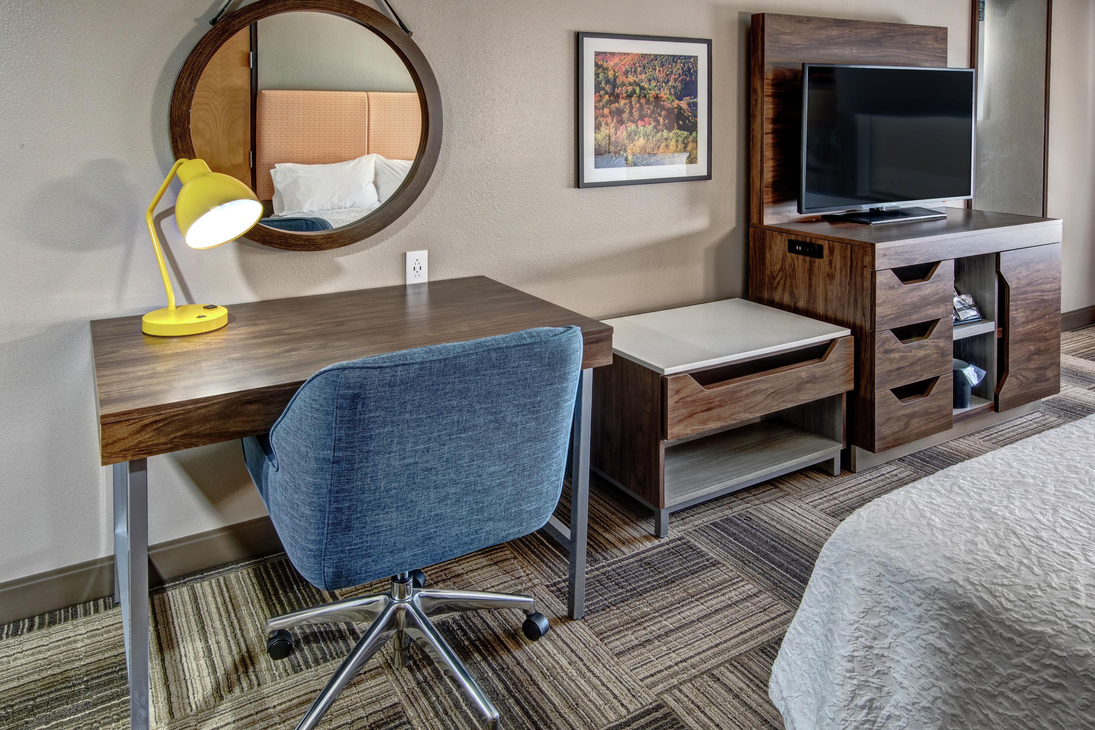 A hotel room with a wooden desk, a blue office chair, a television, a bed, a round mirror, and a framed picture on the wall.