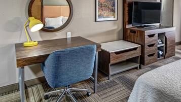 A hotel room with a wooden desk, a blue office chair, a television, a bed, a round mirror, and a framed picture on the wall.