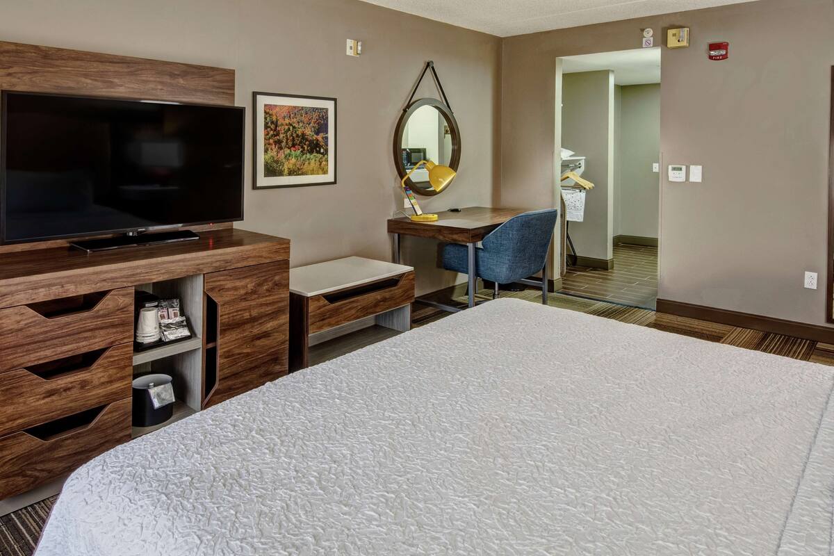 A hotel room with a large bed, a desk with a chair, a television, a mirror, and a small shelf with items.