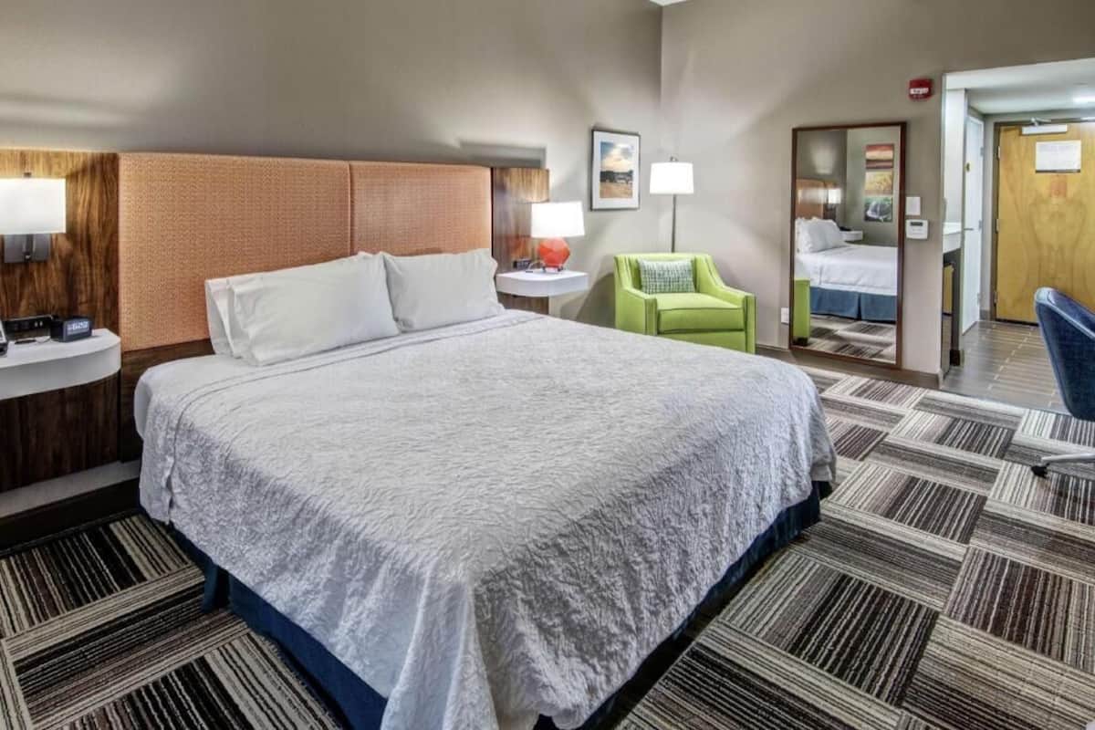 A hotel room with a large bed, a bedside table with a lamp, a green chair, and a mirror.