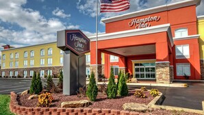 A Hampton Inn by Hilton hotel with a red facade, American flag, and a landscaped entrance area.