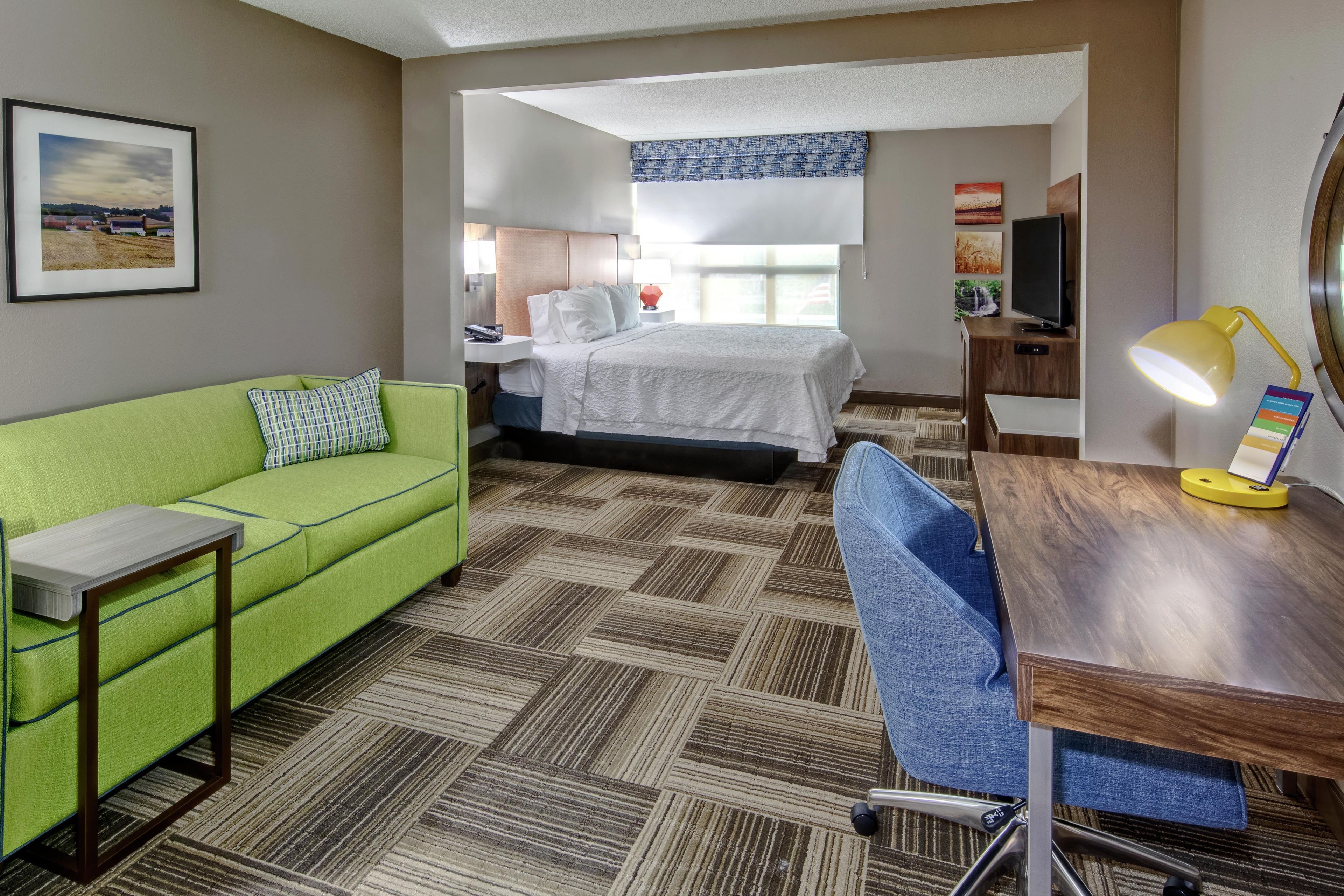 Habitación de hotel con sofá verde, cama con ropa blanca, escritorio con lámpara y televisor.