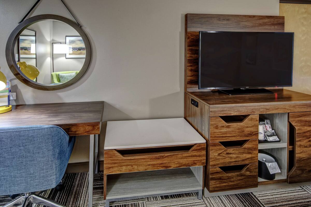 A hotel room with a wooden desk, a flat-screen TV, a mirror, and a small seating area.