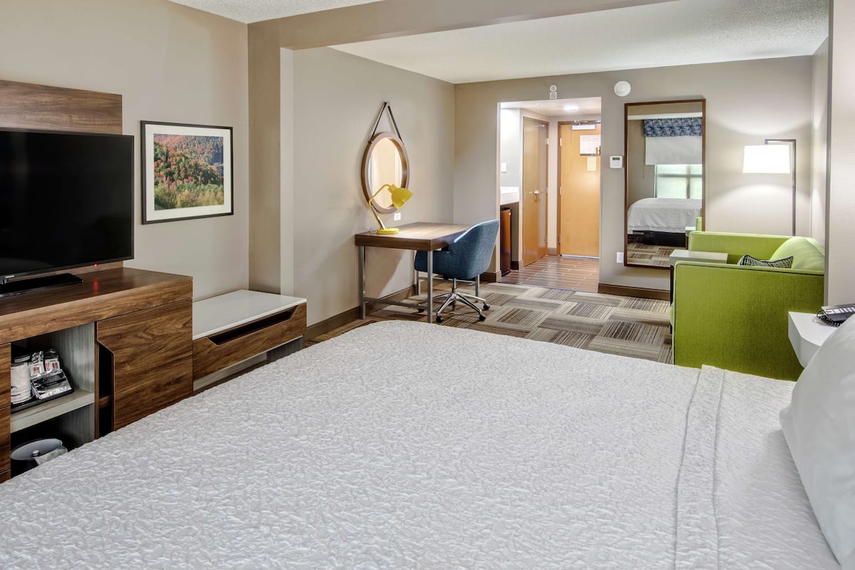 A hotel room with a bed, a desk with a chair, a television, a mirror, and a green sofa.