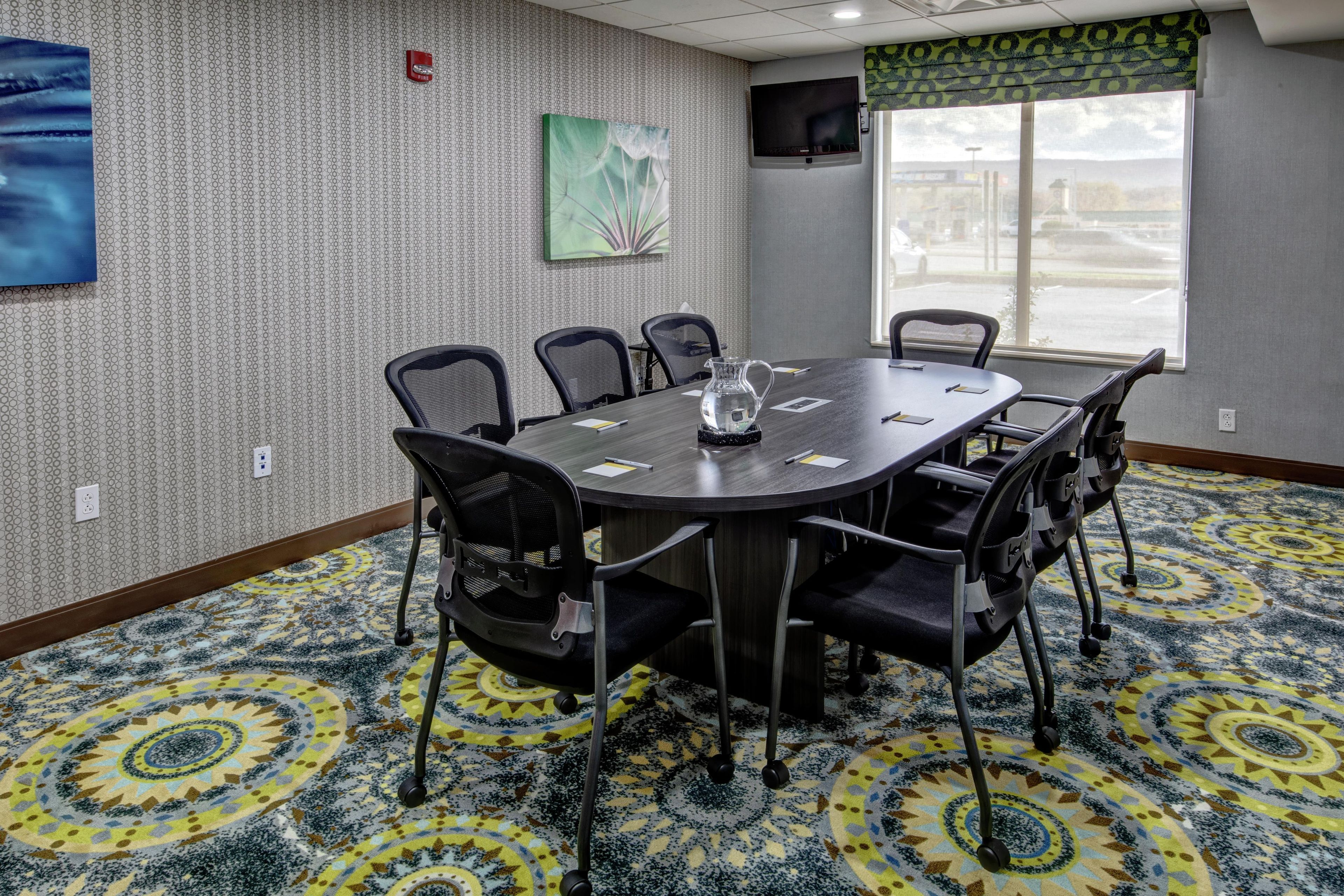 A conference room with a round table, six chairs, a fire alarm, a painting, and a window with a view of the street.