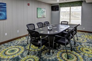 A conference room with a round table, six chairs, a fire alarm, a painting, and a window with a view of the street.