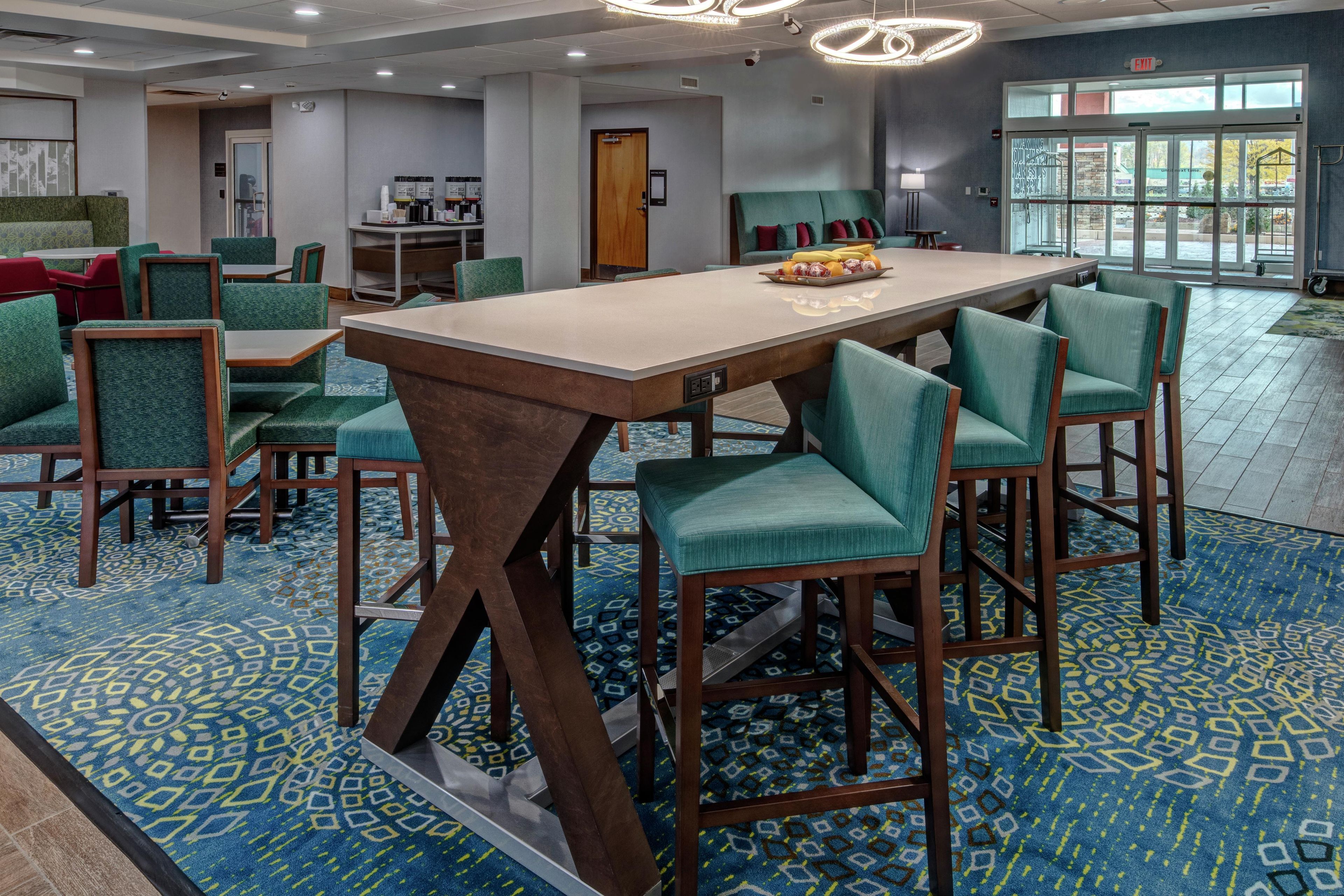 A modern hotel lobby with a central dining table, green upholstered chairs, patterned carpet, and a view through the glass doors.
