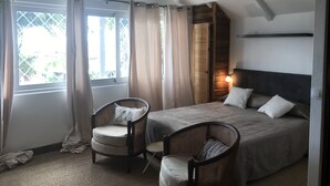 A bedroom with a bed, two wicker chairs, a window with curtains, and a hanging light fixture. - Saint John Perse (Pointe-à-Pitre)