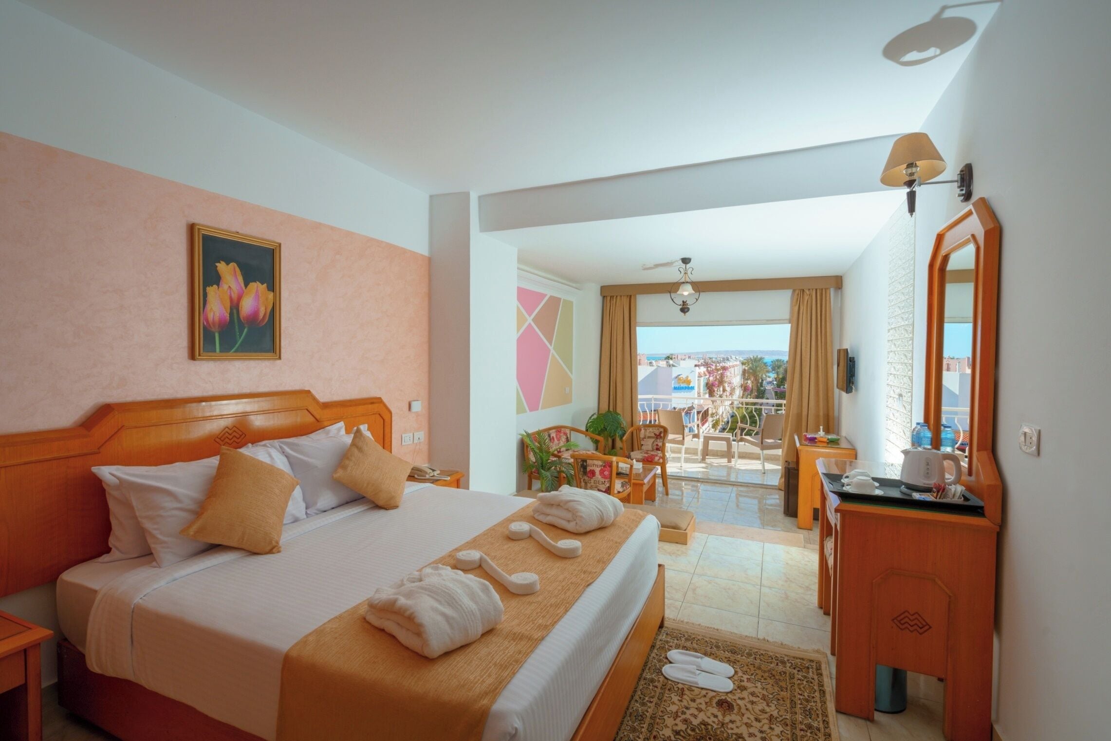A hotel room with a bed, a desk, a chair, a balcony with a view, and a painting on the wall.