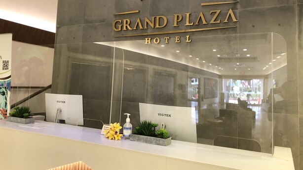 A reception desk at Grand Plaza Hotel with a modern design, a clear glass partition, and a sign indicating the hotel name. - Grand Plaza Hotel (Tamuning)
