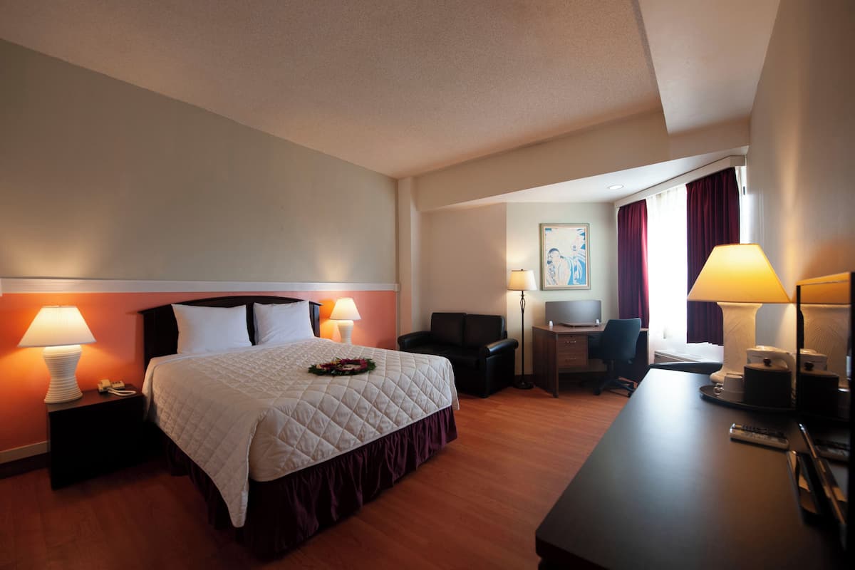 Quarto de hotel com uma cama grande, duas luminárias de cabeceira, uma mesa com cadeira, uma mesinha e uma janela com cortinas.