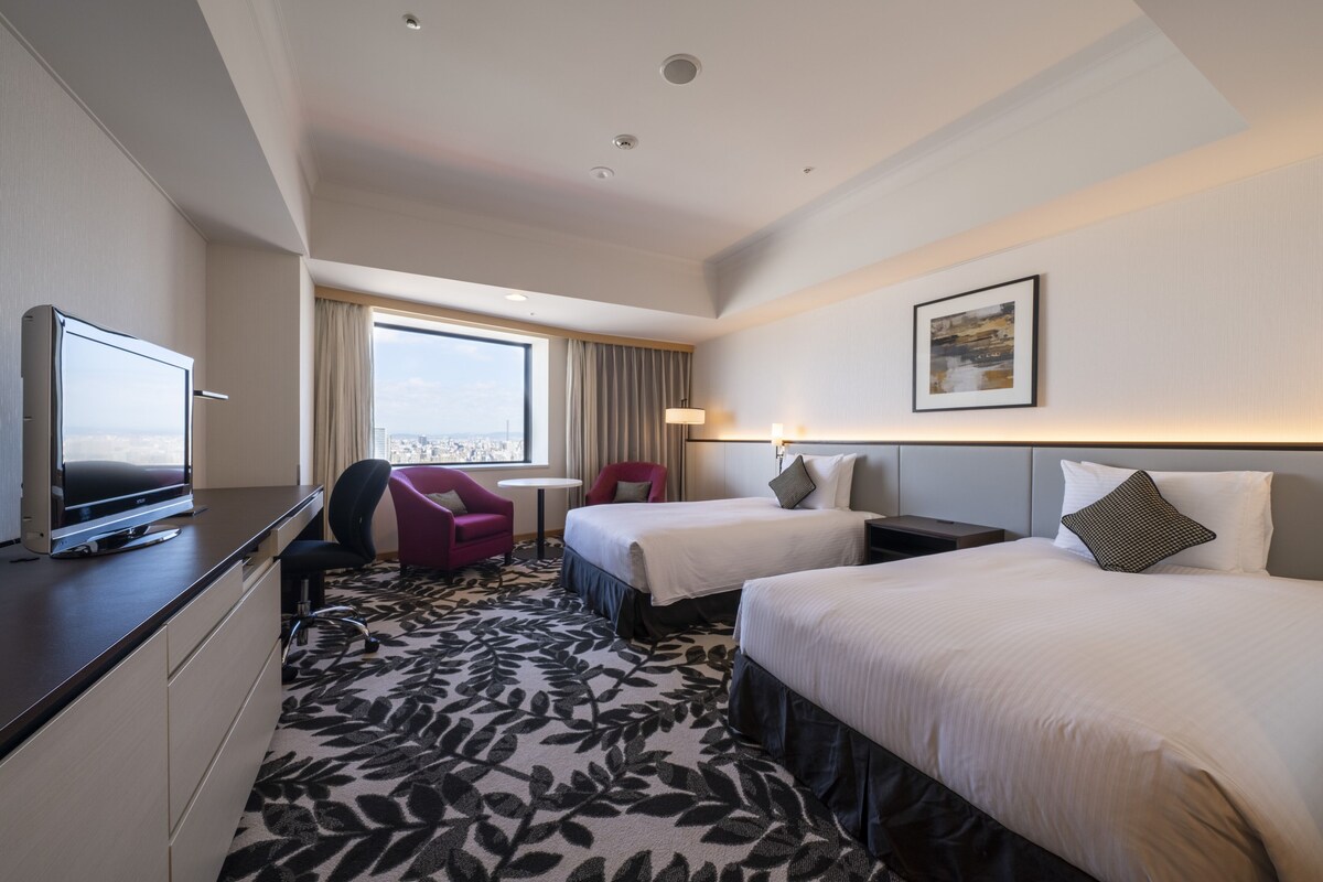 Habitación de hotel con dos camas, un escritorio, una silla, un televisor y una ventana con vistas.