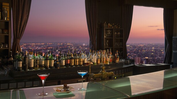 A bar with a city view at sunset, featuring a variety of bottles and two martini glasses.