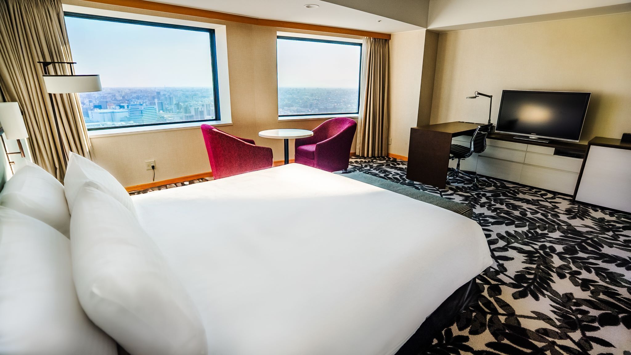 Habitación de hotel con una cama grande, dos ventanas con vista a la ciudad, una pequeña mesa redonda, una silla roja, un escritorio con un televisor y una alfombra estampada.