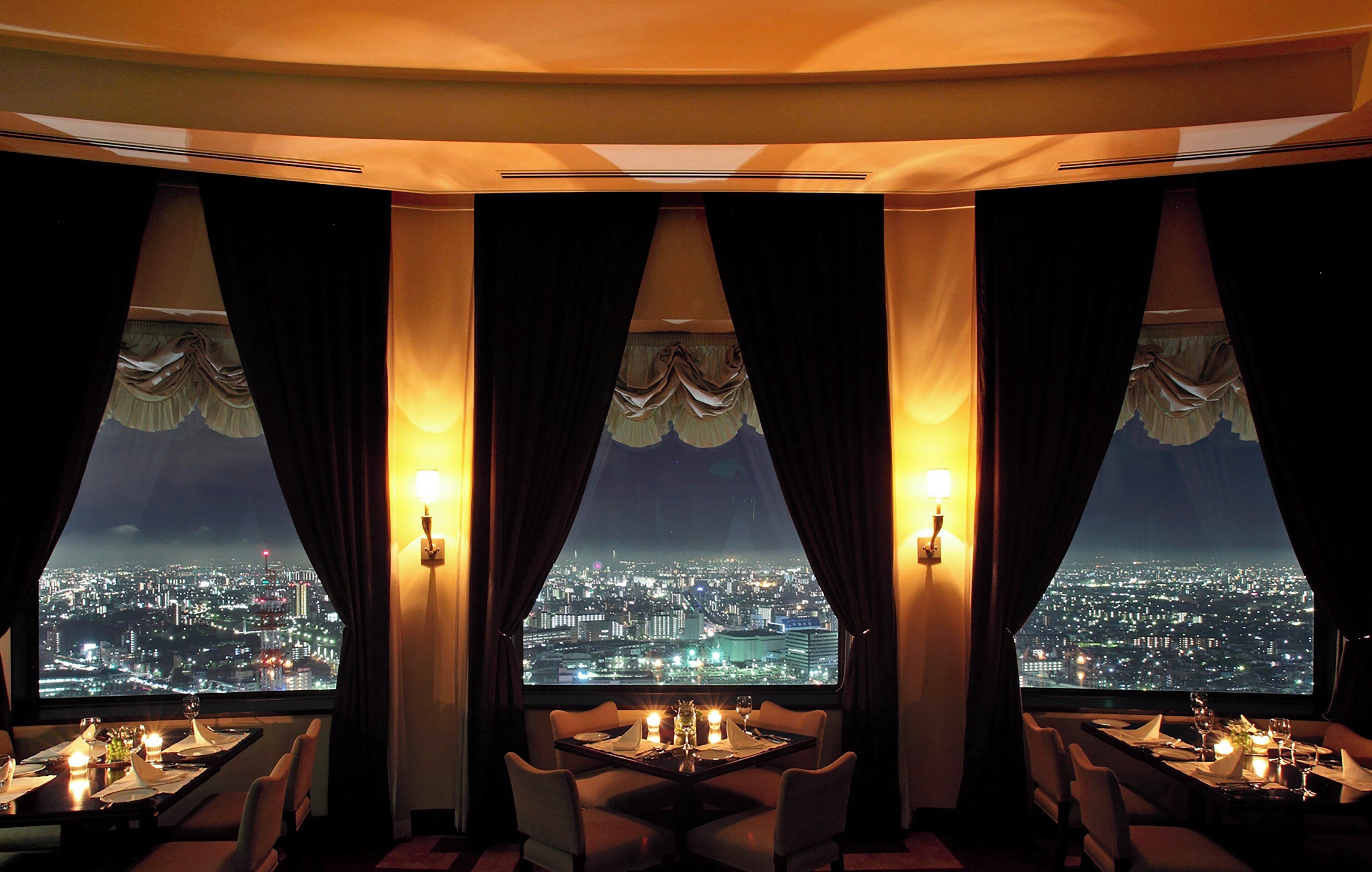 A dining area with a city view, featuring tables set with candles, chairs, and curtains.
