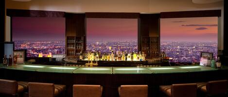 A bar with a cityscape view, bar stools, and a well-lit ceiling.