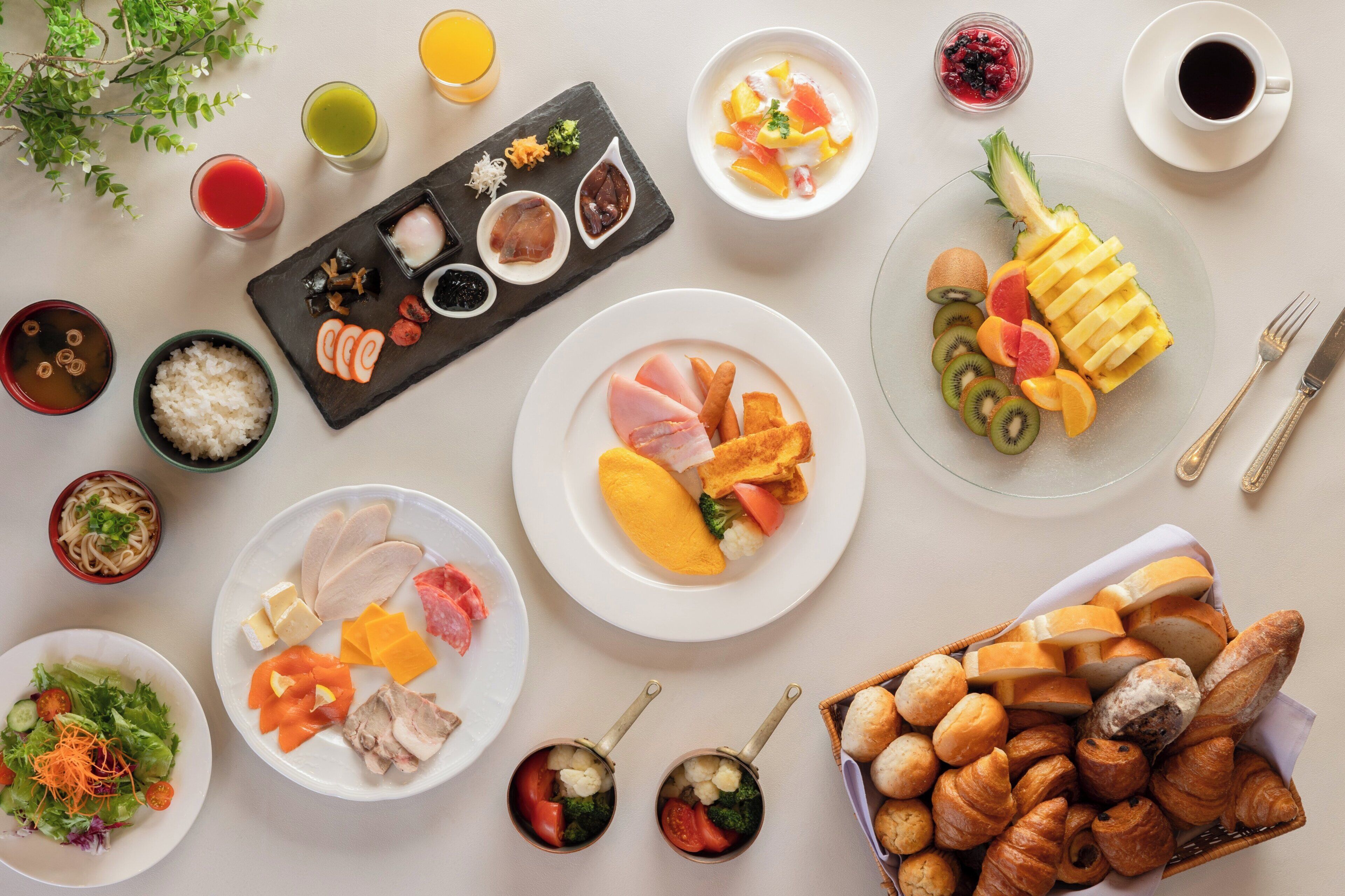 A variety of dishes and drinks on a table, including plates with sliced fruits, vegetables, and meats, as well as bowls of soup and rice.