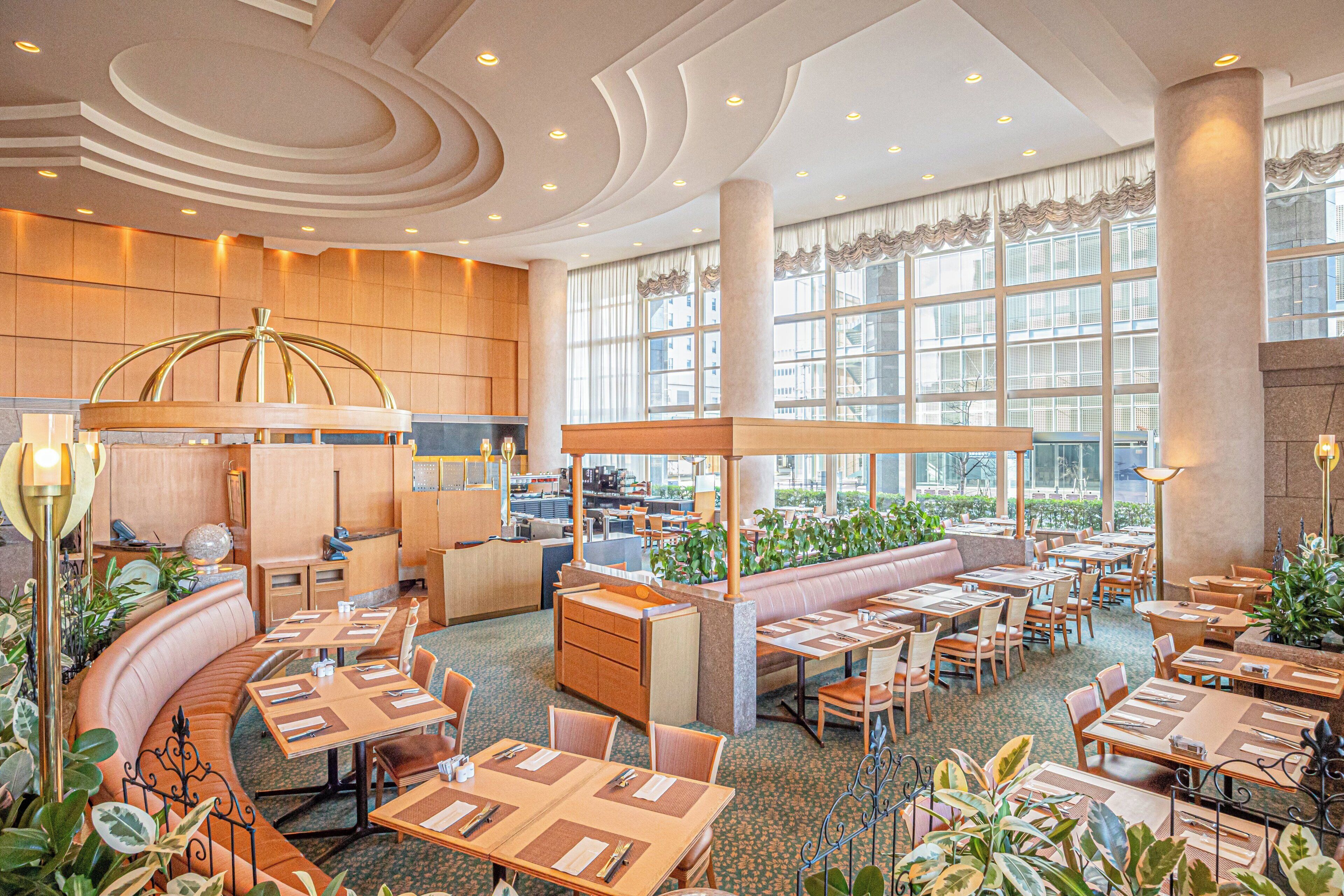 Un amplio vestíbulo de hotel con una zona de estar central, varias mesas de comedor y grandes ventanales que ofrecen luz natural.
