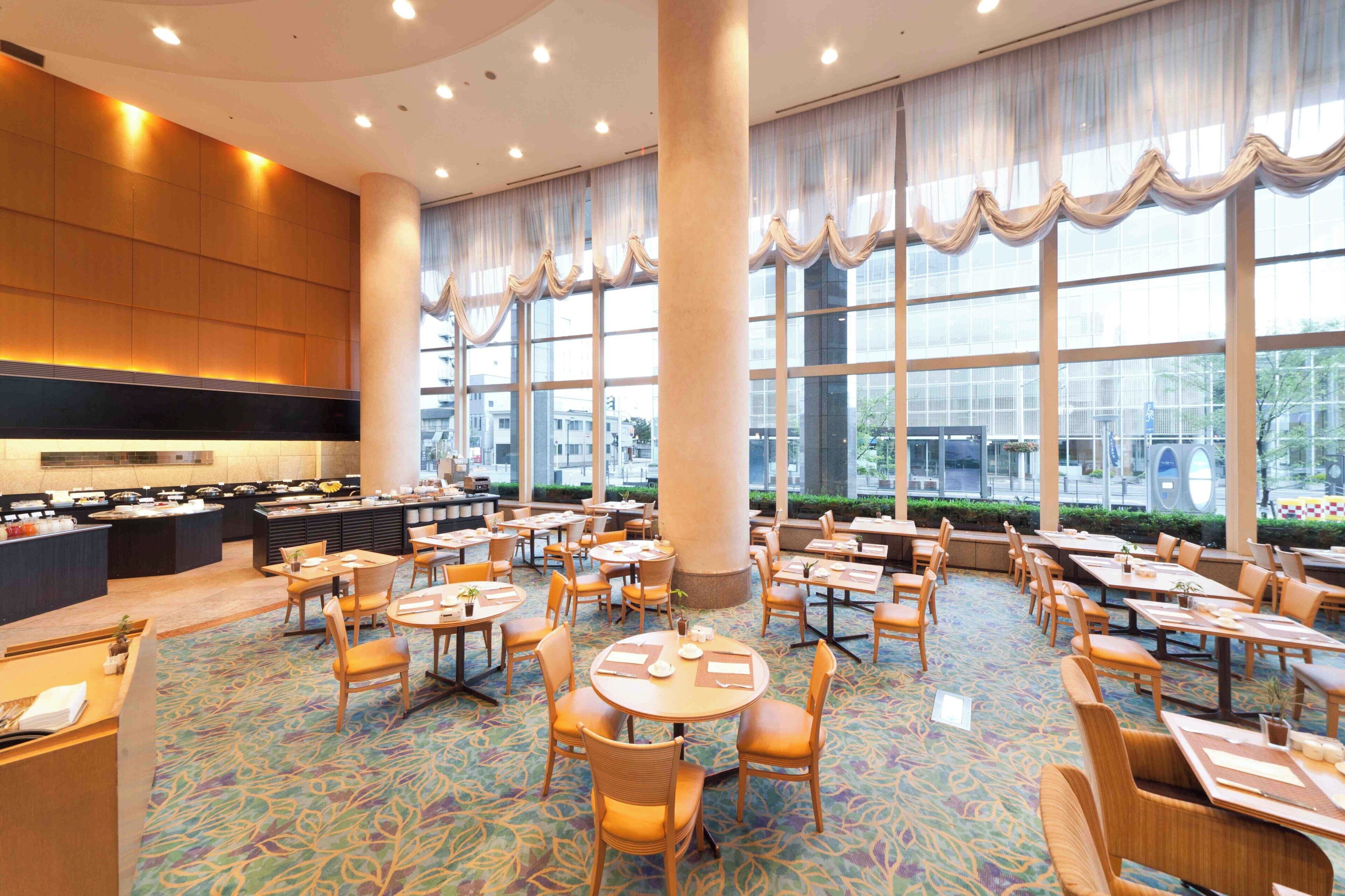 A hotel dining area with round tables, chairs, a buffet, and large windows.
