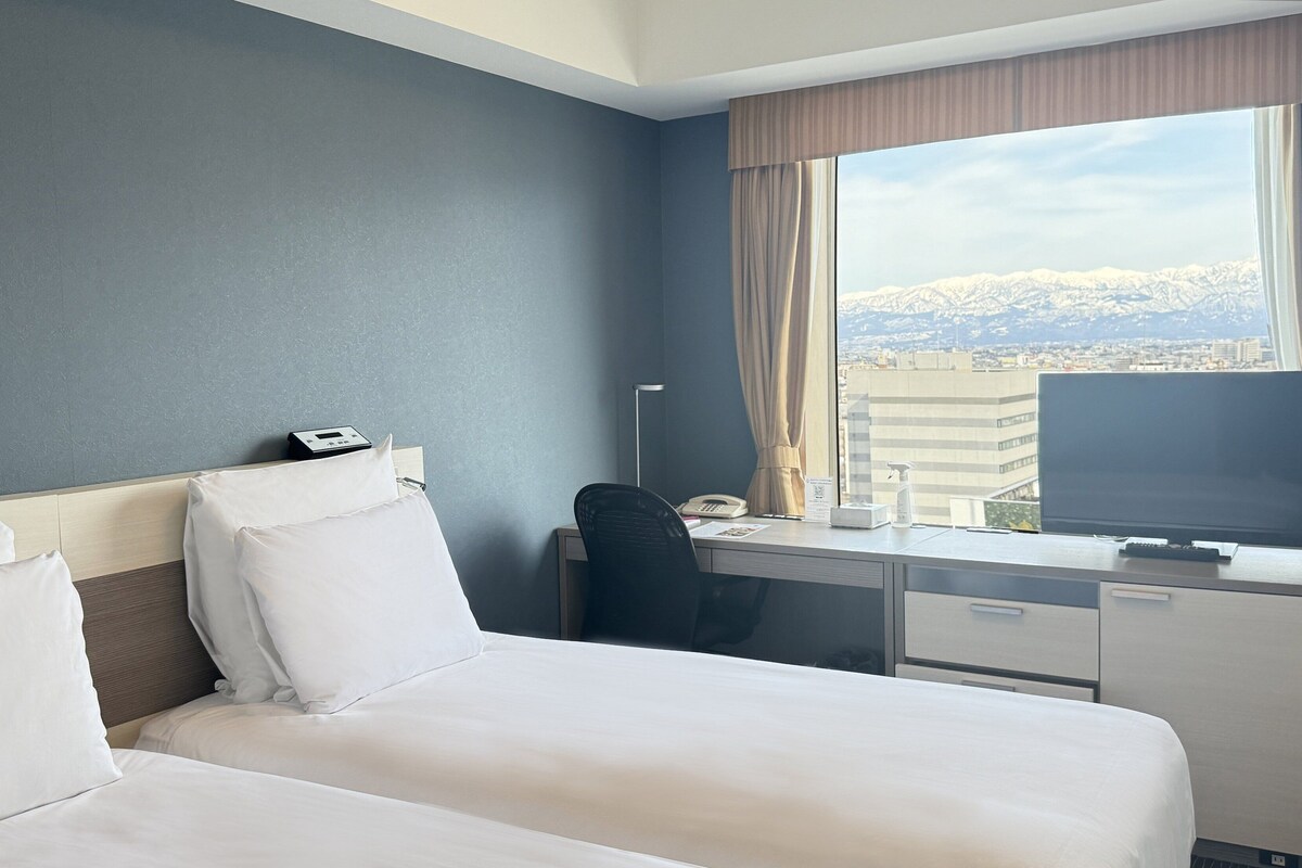 A hotel room with a bed, desk, chair, and a large window with a cityscape view.