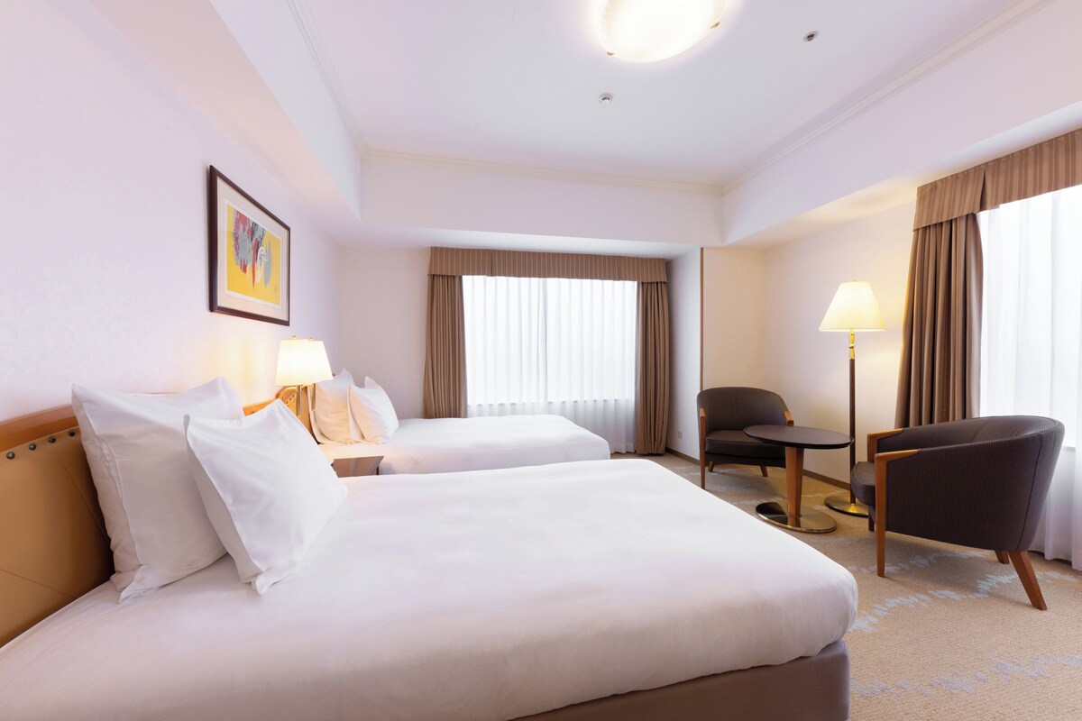 A hotel room with two beds, a chair, a table, and a lamp.