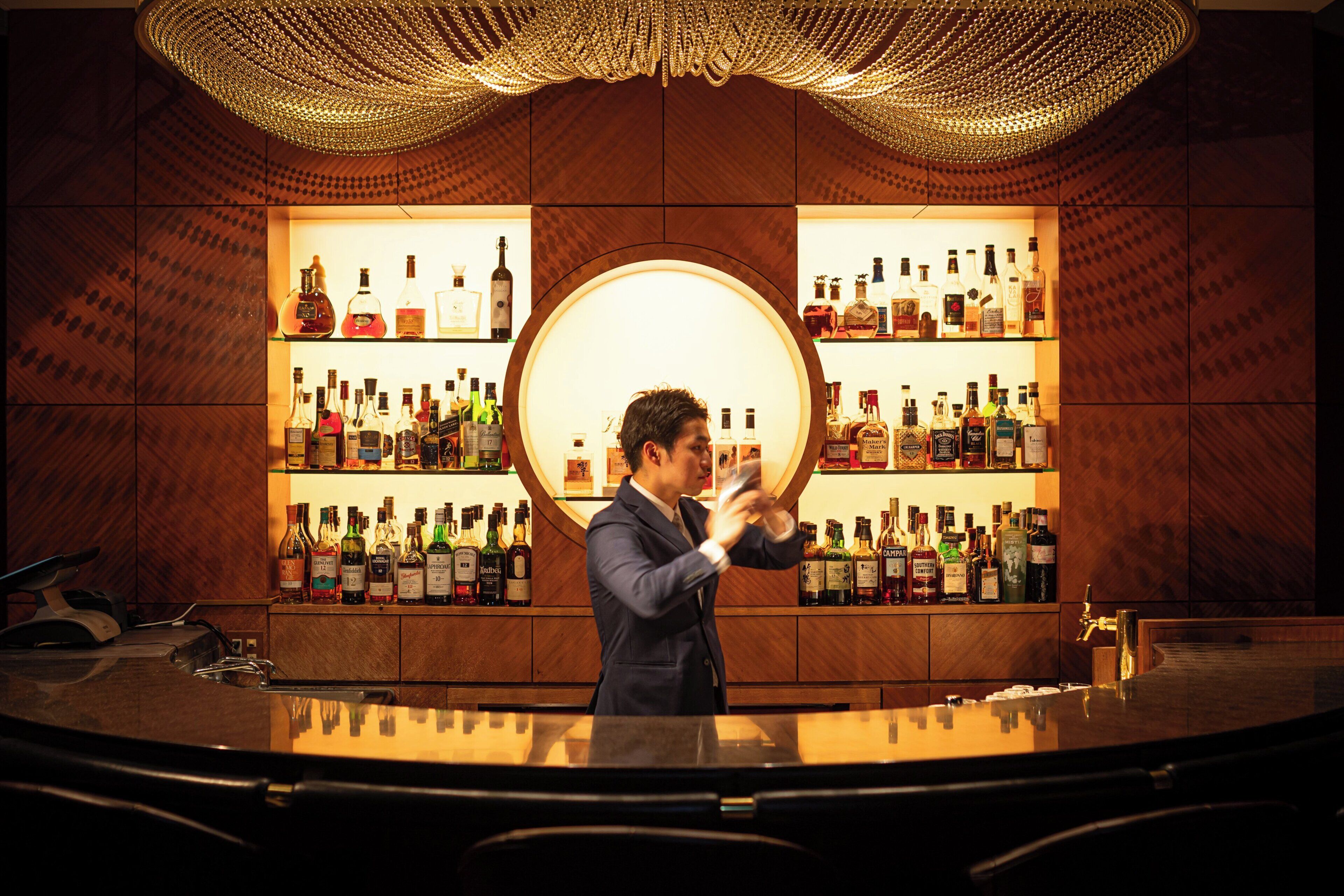 A bar area with a circular light fixture, shelves of liquor bottles, and a bar counter.