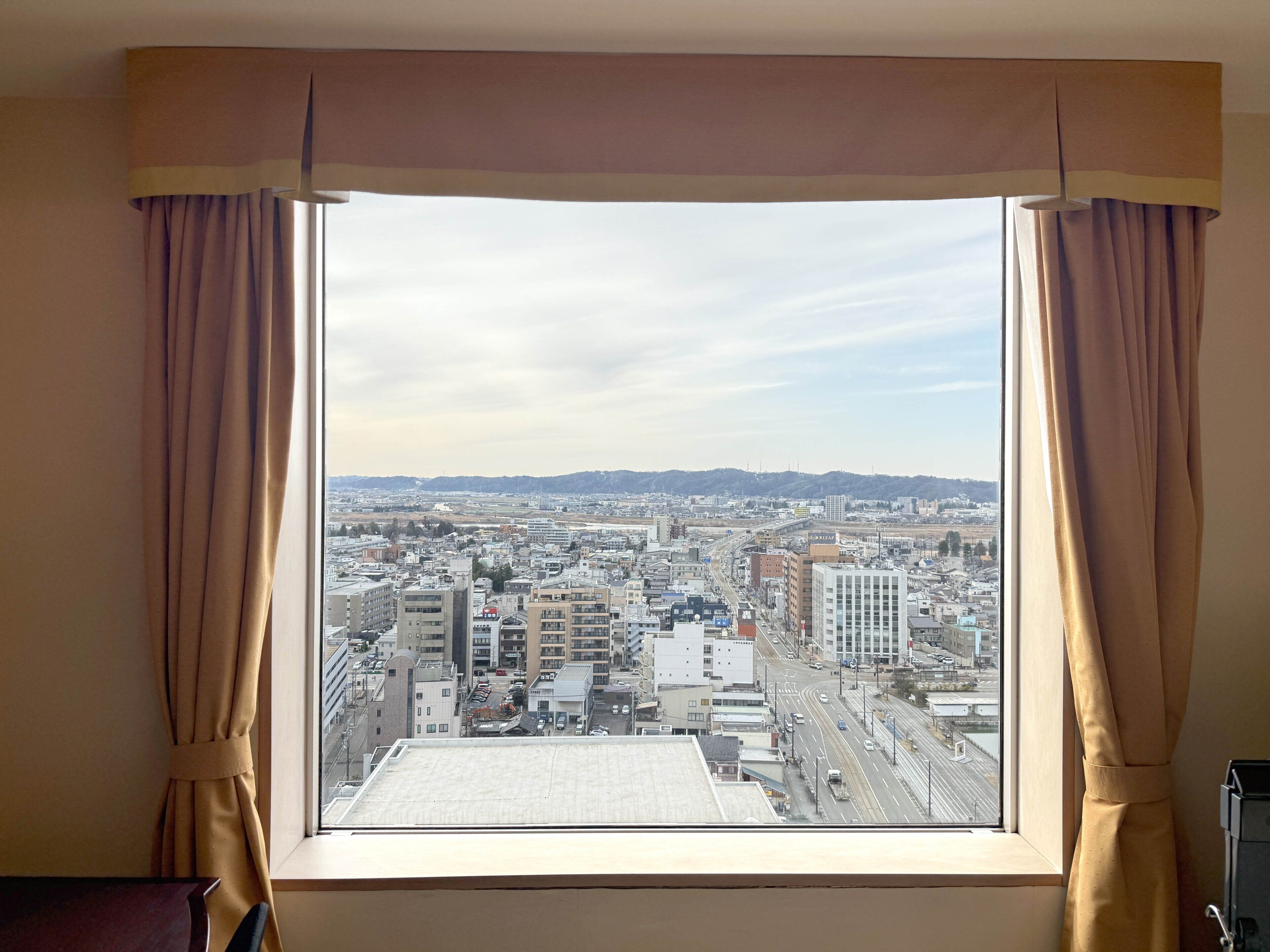 A window with curtains, a view of a cityscape, and a building with a flat roof.