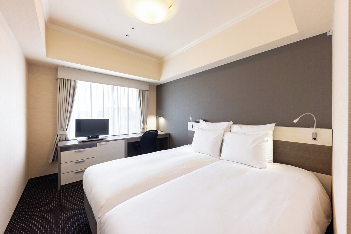 A hotel room with a bed, a desk with a TV, a chair, and a window with curtains.
