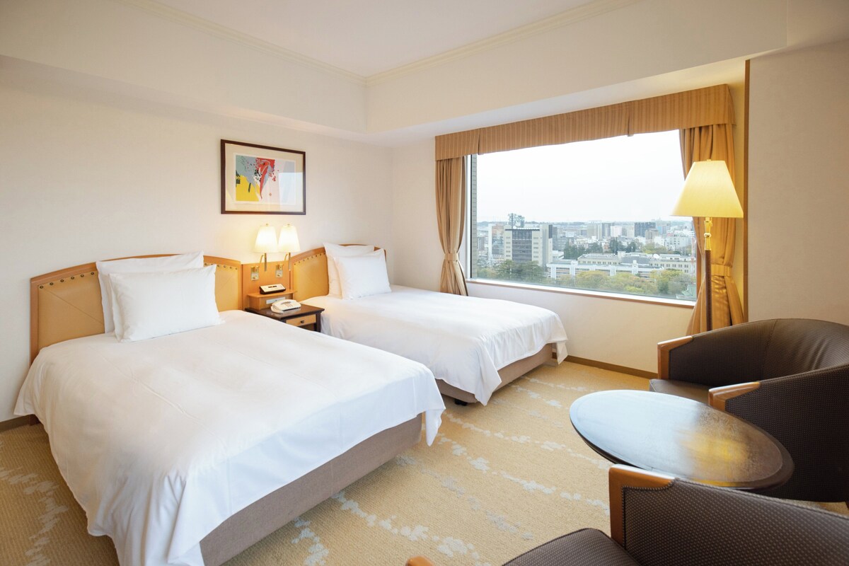 A hotel room with two beds, a desk, a chair, and a large window with a city view.