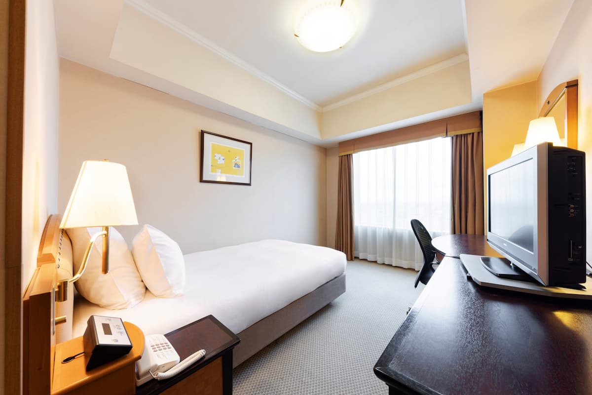 Una habitación de hotel con una cama, una mesita de noche con lámpara, un escritorio con computadora, un televisor y una ventana con cortinas.