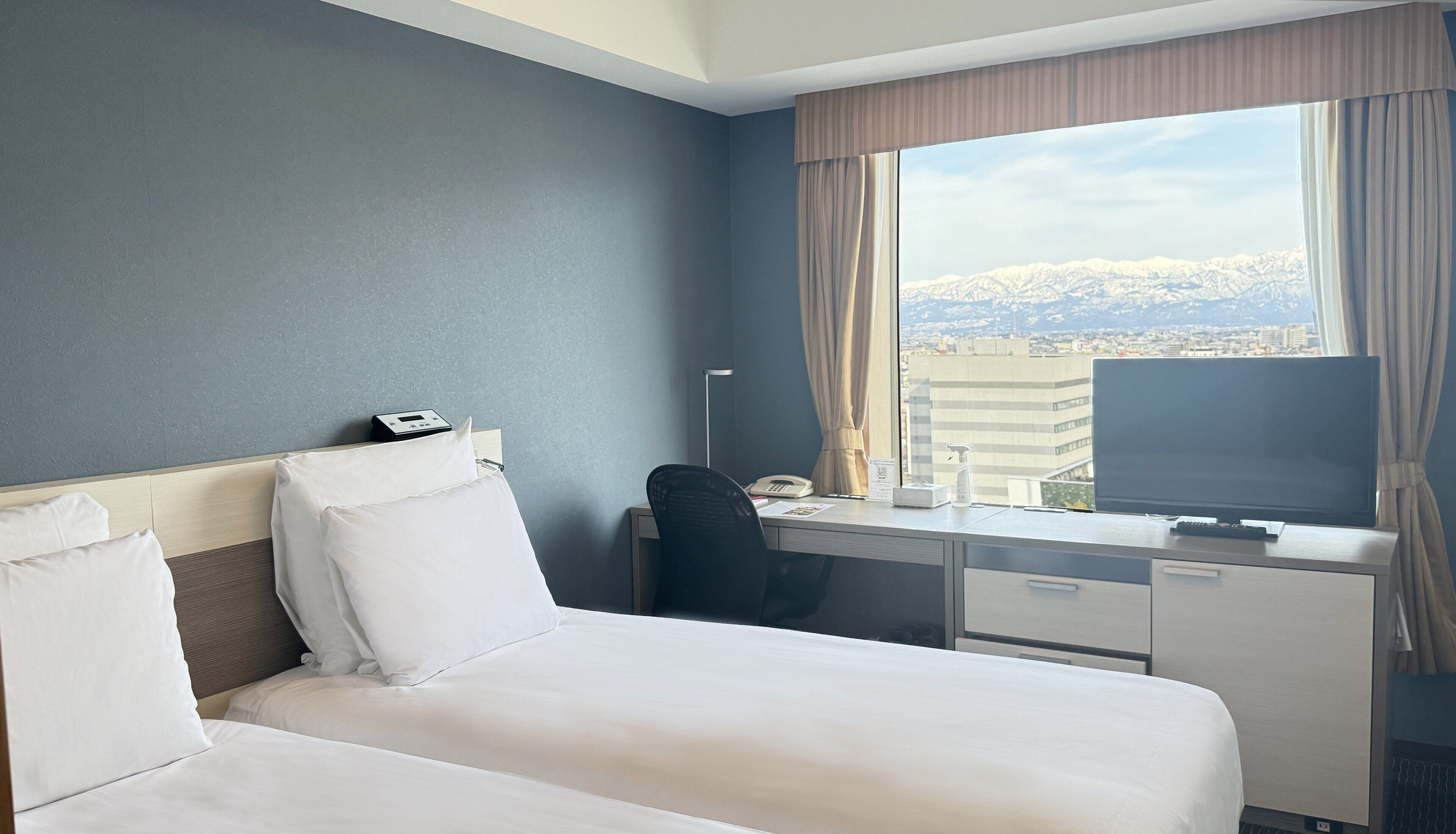 A hotel room with a bed, desk, chair, and a large window with a cityscape view.