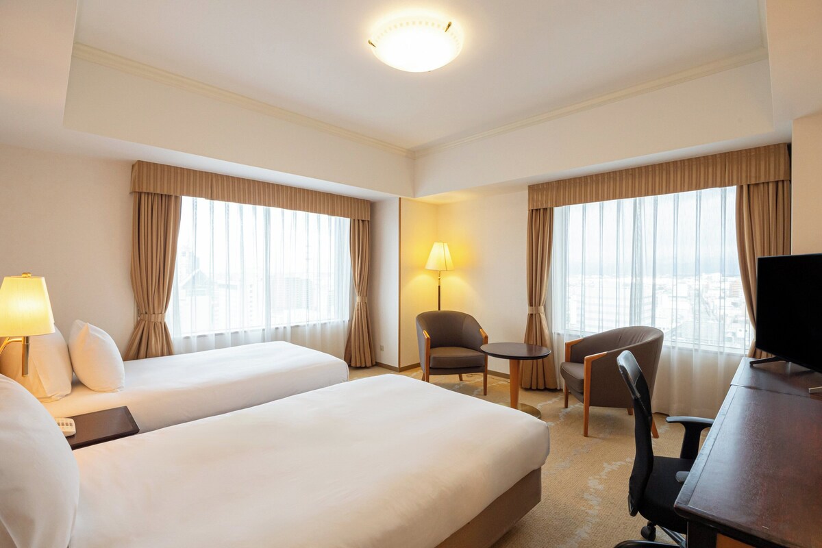 A hotel room with two beds, a desk, a chair, a TV, and a lamp.