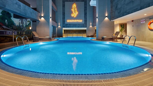 A large indoor swimming pool with a modern design, surrounded by lounge areas and a prominent hotel logo. - Mardhiyyah Hotel and Suites (Shah Alam)