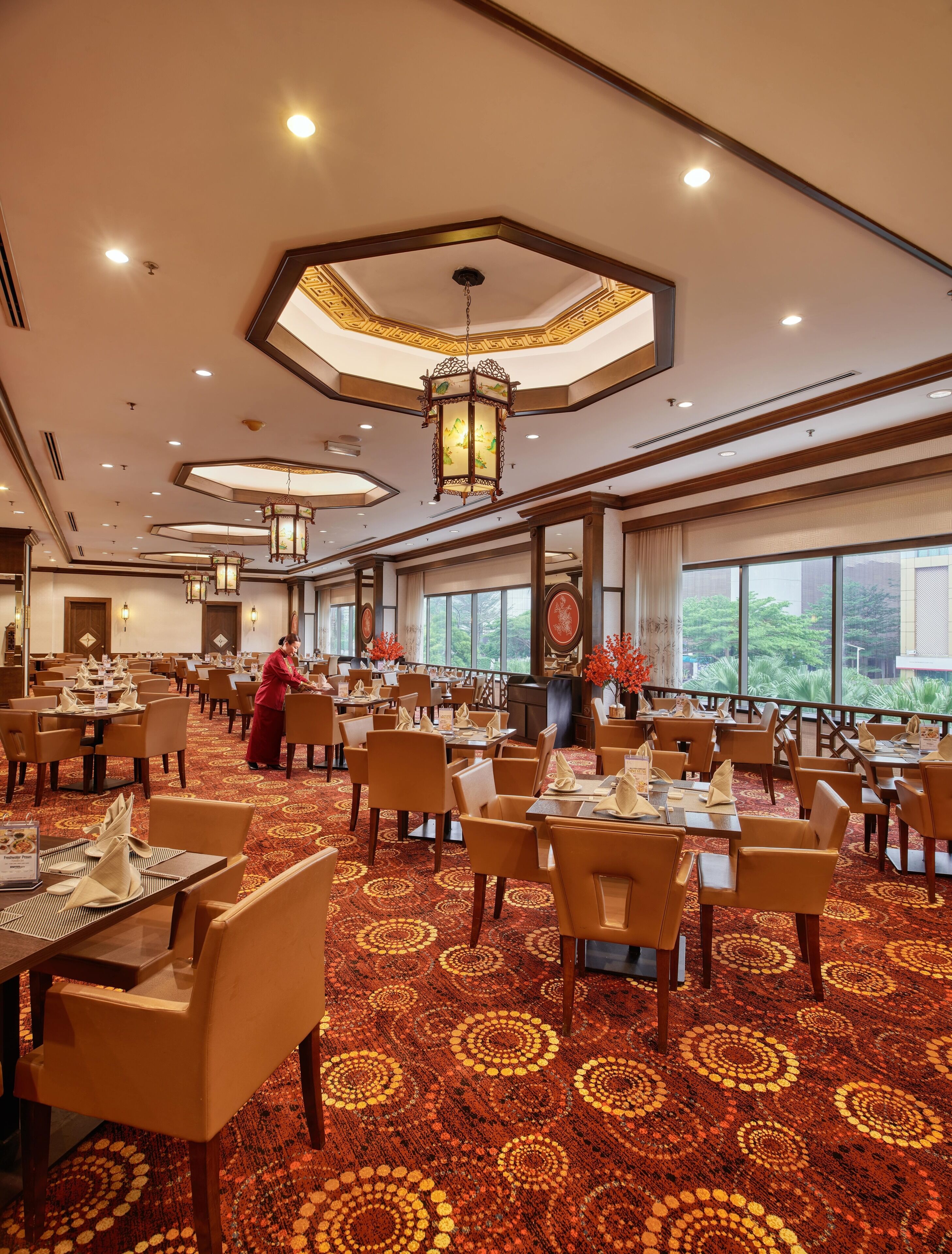 A spacious dining area with multiple tables and chairs, a patterned carpet, and a view of greenery outside.
