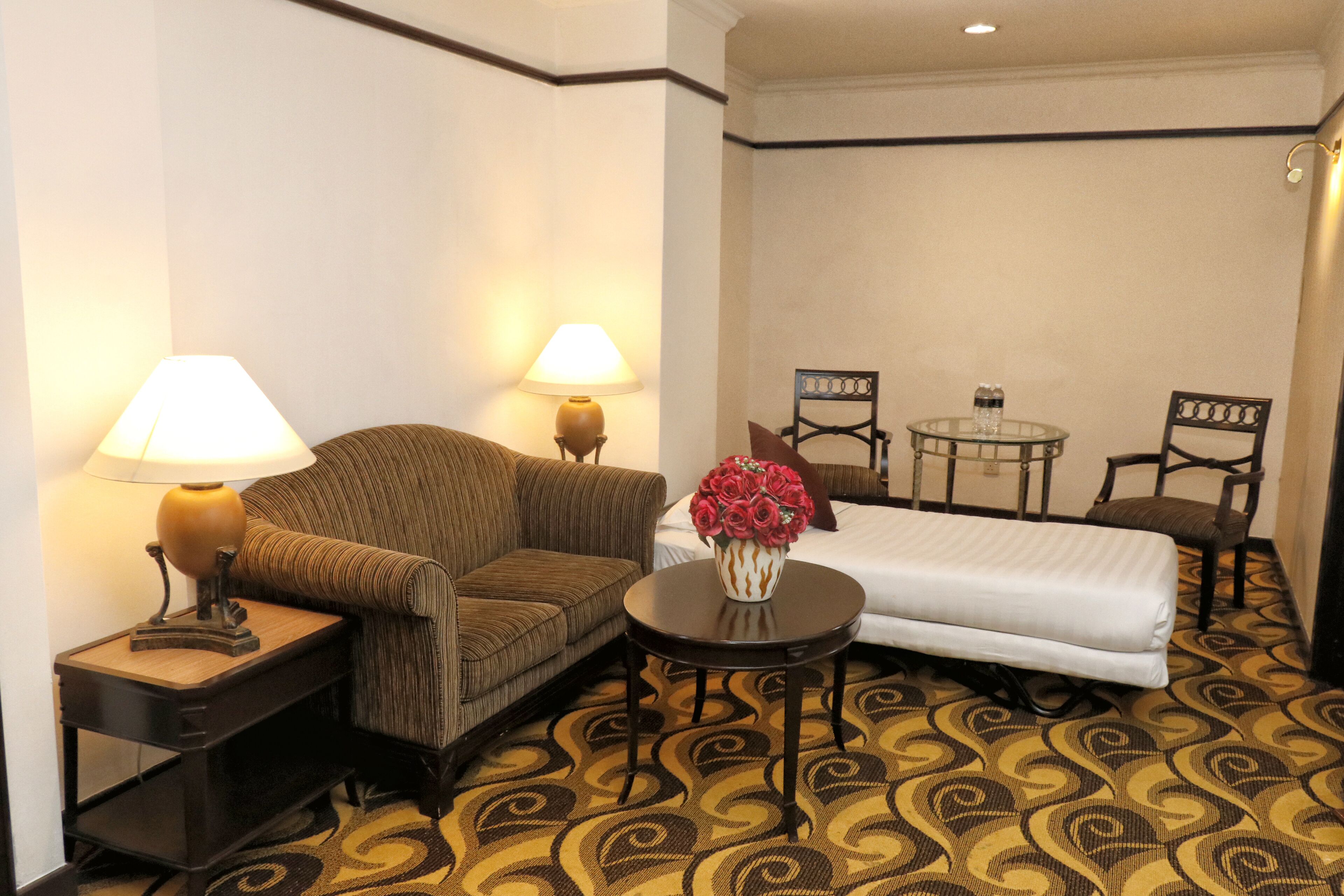 Una habitación de hotel con sofá, dos sillones, una mesita con un jarrón de flores y una cama.