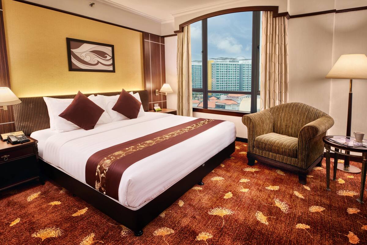 Ein Hotelzimmer mit einem großen Bett, einem Sessel, einem Nachttisch, einer Lampe und Blick auf Gebäude durch ein großes Fenster.